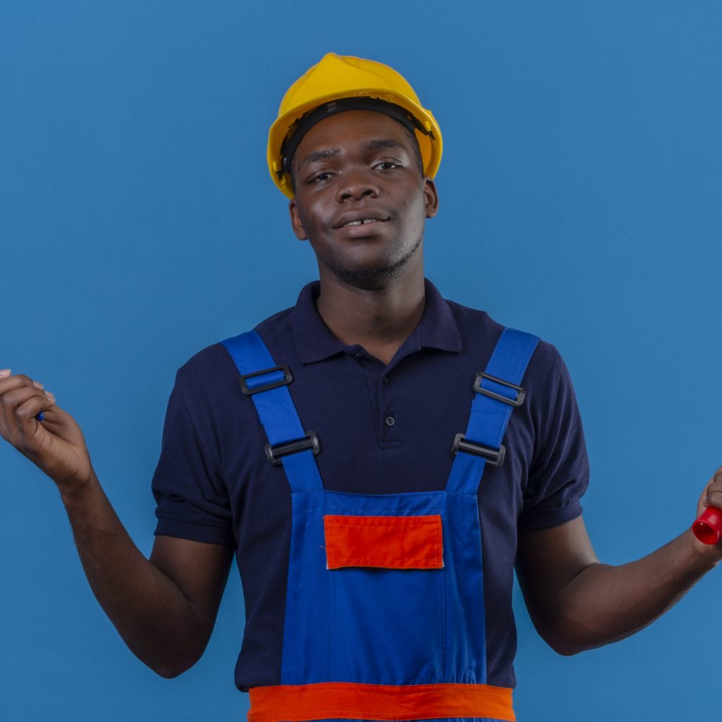young african american builder man wearing construction uniform and safety helmet holding brush and paint roller looking at camera smiling friendly standing over blue background